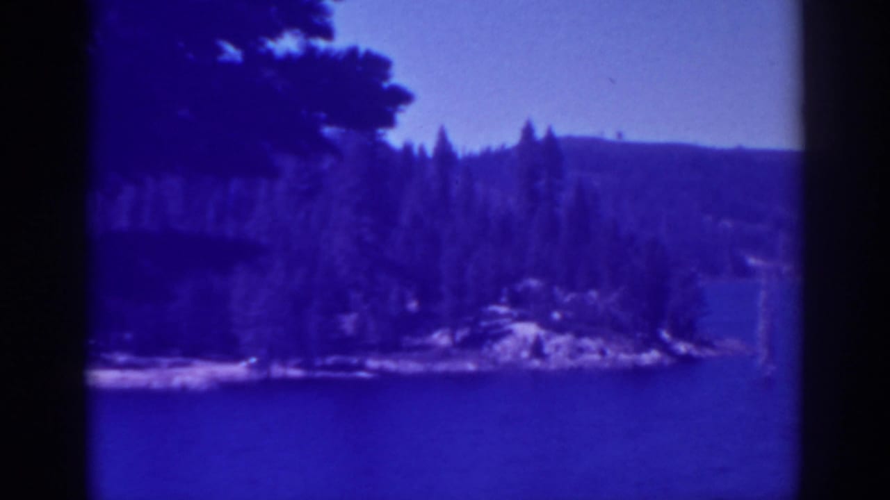 Blue lake water with evergreen shoreline, rocky point, distant hills, and clear sky.