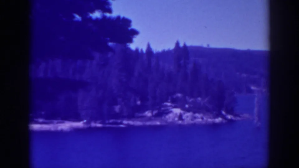 Blue-tinted 1938 Bucks Lake view across open water toward evergreen trees, a rocky shoreline point, low hills, and clear sky.