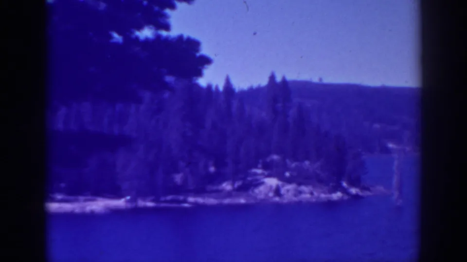 1938 Bucks Lake home-movie frame showing blue water in the foreground, evergreen trees along the shore, and hazy hills beyond.