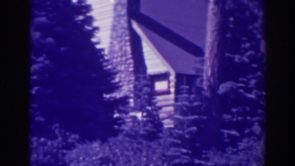 1938 Bucks Lake 8mm frame showing a log cabin, steep roof, stone chimney, pine trunks, and dense evergreen foliage.