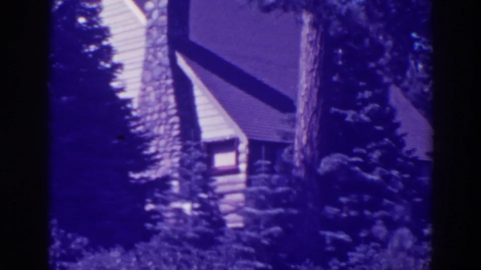 Bucks Lake cabin exterior in a purple-tinted 8mm frame, with a stone chimney and roof partly obscured by pines.