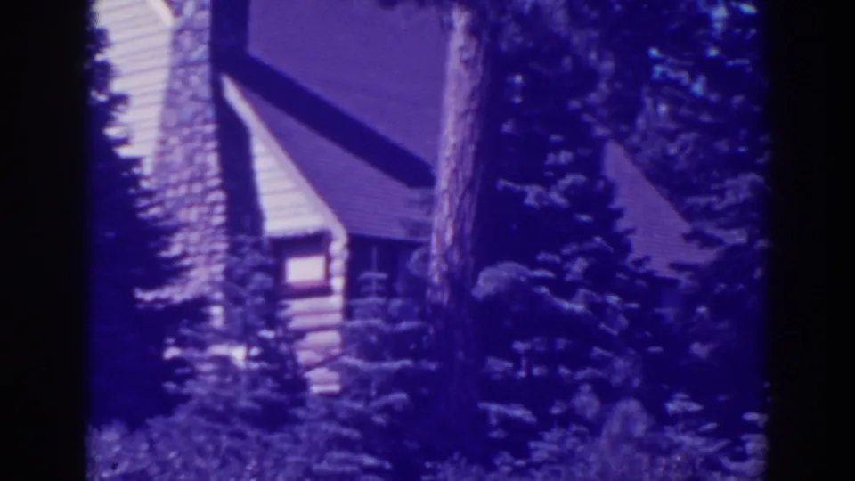 Purple-blue 8mm frame of the Bucks Lake log cabin facade, with a steep roofline, chimney, pines, and brush.