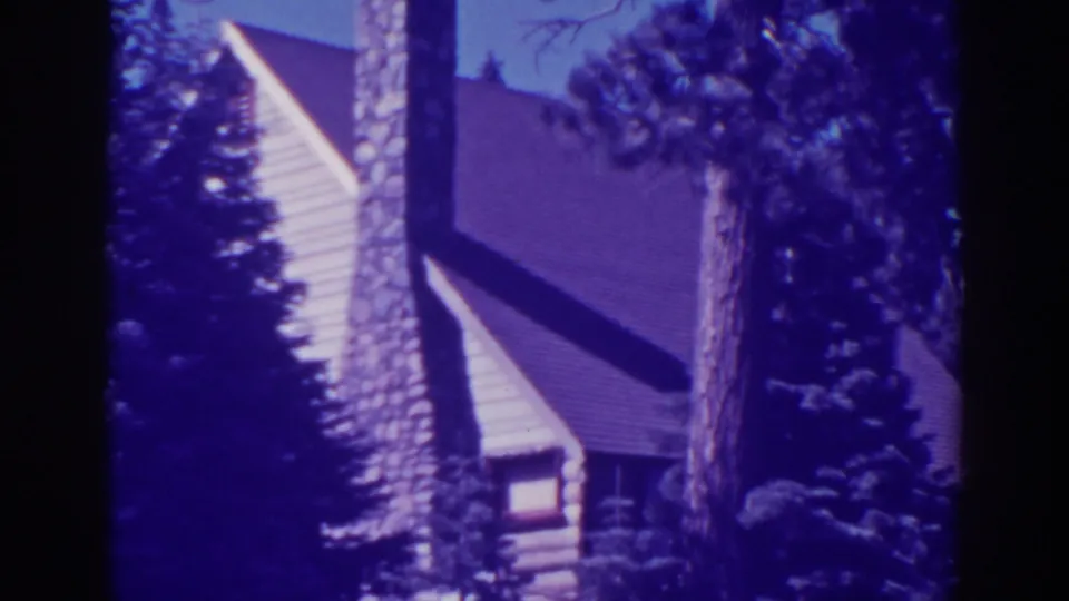 Bucks Lake cabin exterior in 1938 footage, with a stone chimney rising beside the roof and trees partially obscuring the siding.
