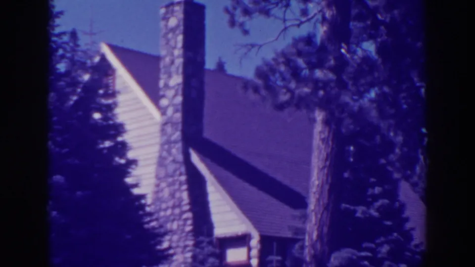 Frame from the 1938 Bucks Lake clip showing the cabin side, chimney stones, roofline, window area, and surrounding pines.
