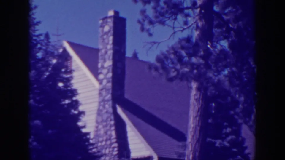 Late frame of the 1938 Bucks Lake cabin view, showing the stone chimney, sloped roof, wood siding, and nearby pine trunks.