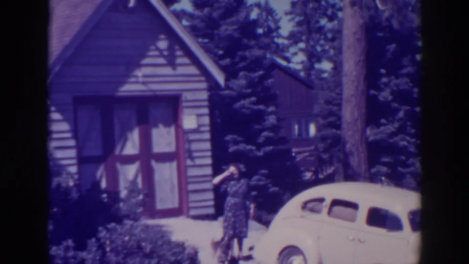 In the 1938 Bucks Lake footage, the woman appears to shade her eyes or look around beside the car, with the cabin and evergreens behind her.