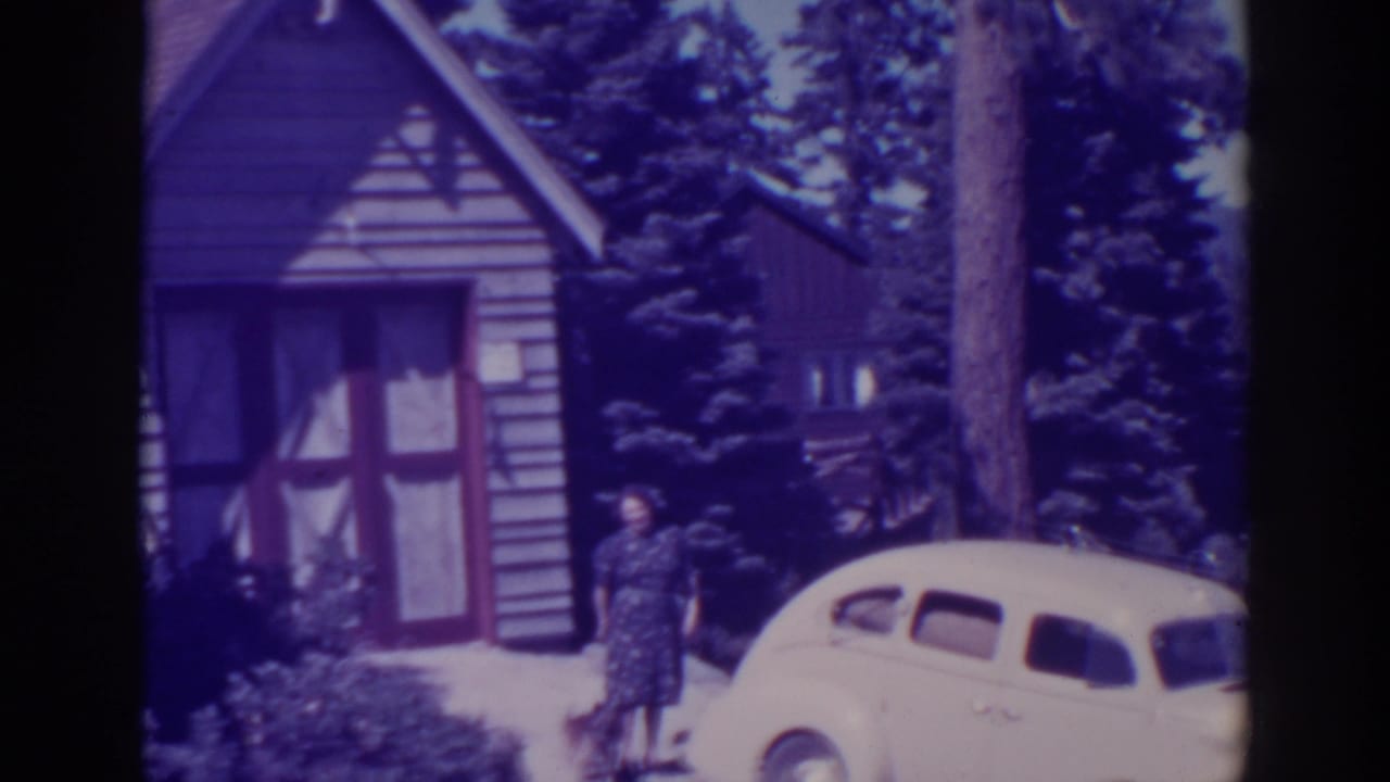 At Bucks Lake, California in supplied 1938 footage, a woman poses near a pale vintage car outside a small cabin surrounded by pines.