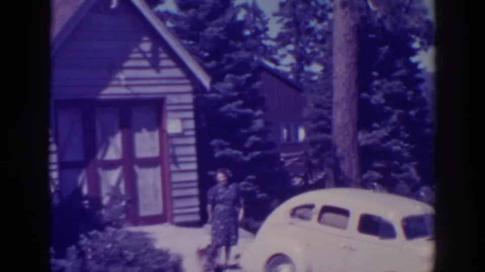 At Bucks Lake, California in supplied 1938 footage, a woman poses near a pale vintage car outside a small cabin surrounded by pines.