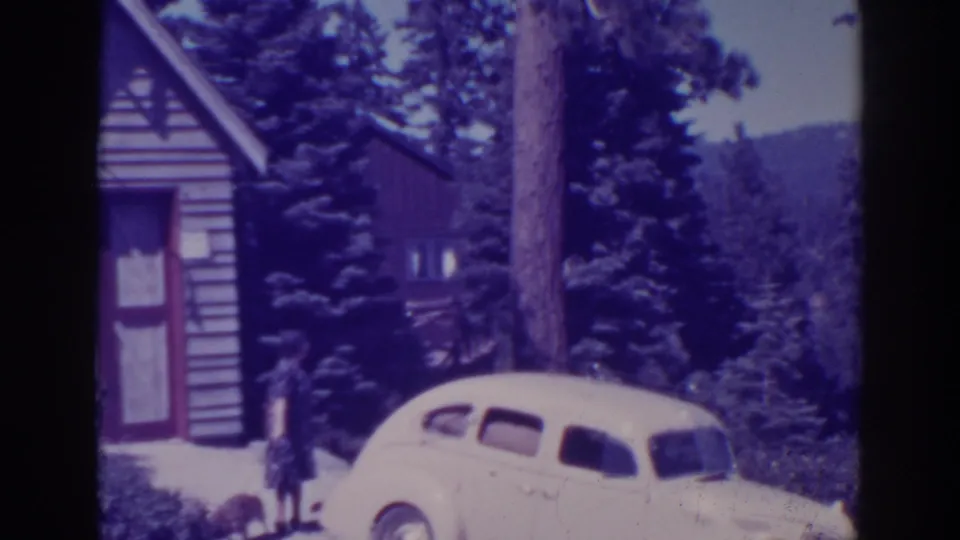 The woman remains near the vintage car and cabin as the wooded Bucks Lake setting and distant hillside become more visible.