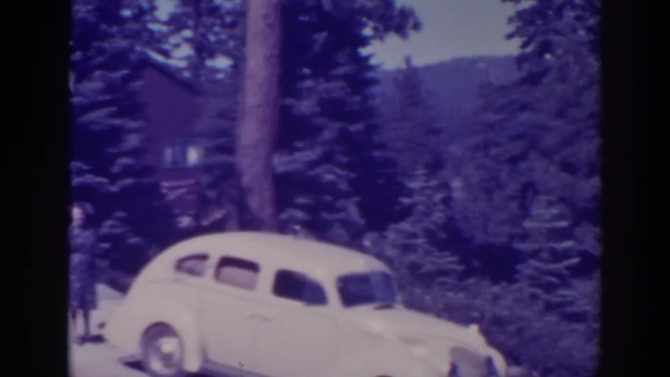 The camera shifts toward the car and forested mountain backdrop, with only part of the cabin and woman visible at the edge.