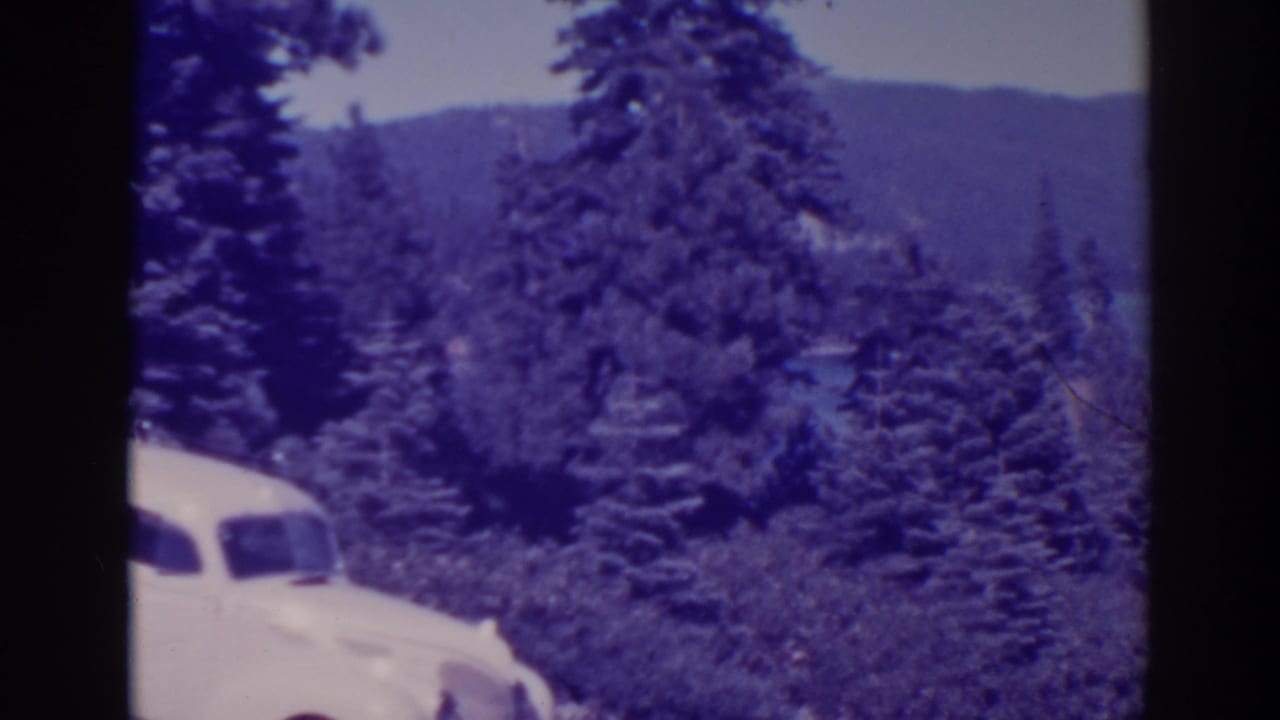 Partial white sedan at left with pine trees, lake water, and hills behind.