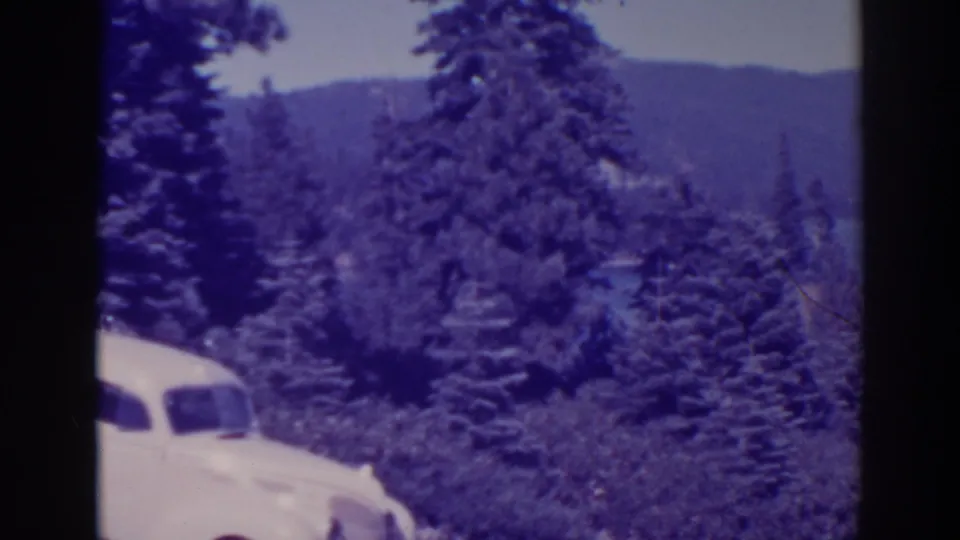White 1930s-era sedan partly visible at Bucks Lake, backed by pines, blue water, and distant hills.