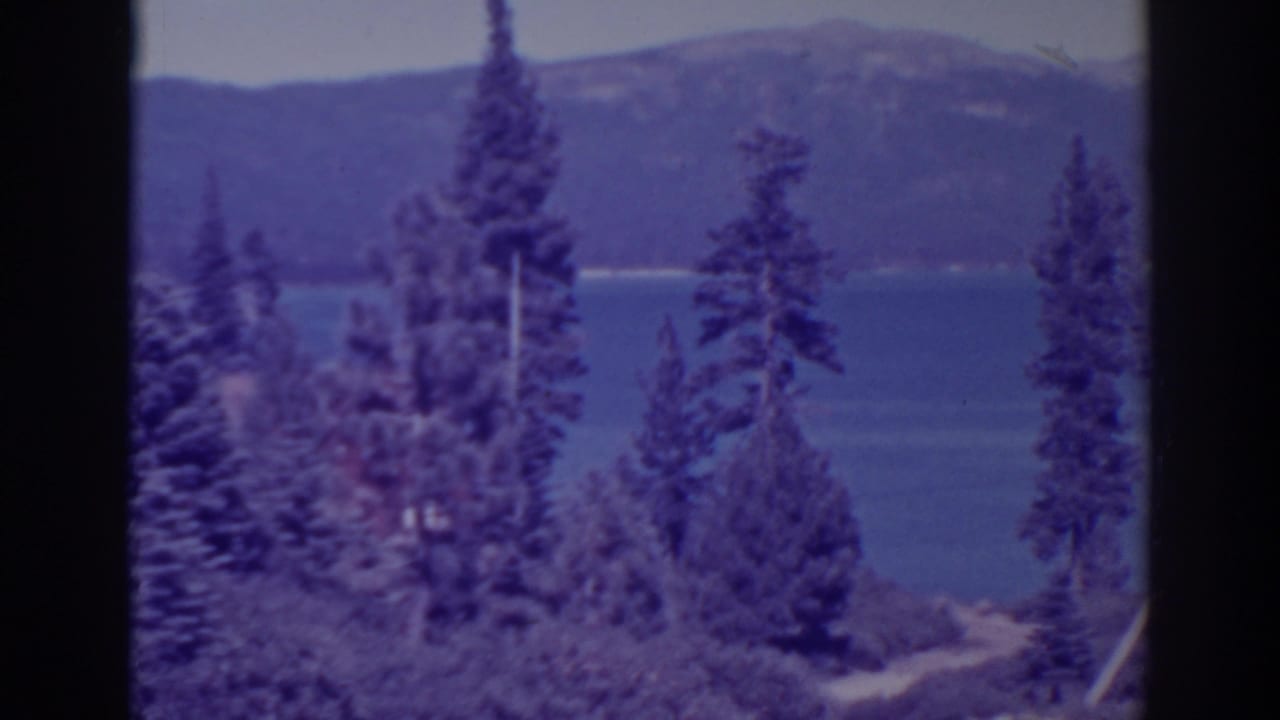 Pine trees frame a blue lake with distant mountains behind it.