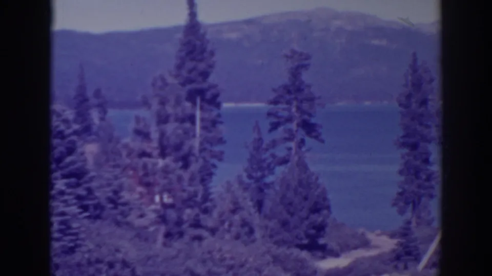 1938 8mm view at Bucks Lake, California, with tall pines in the foreground, blue water, and a distant mountain ridge.