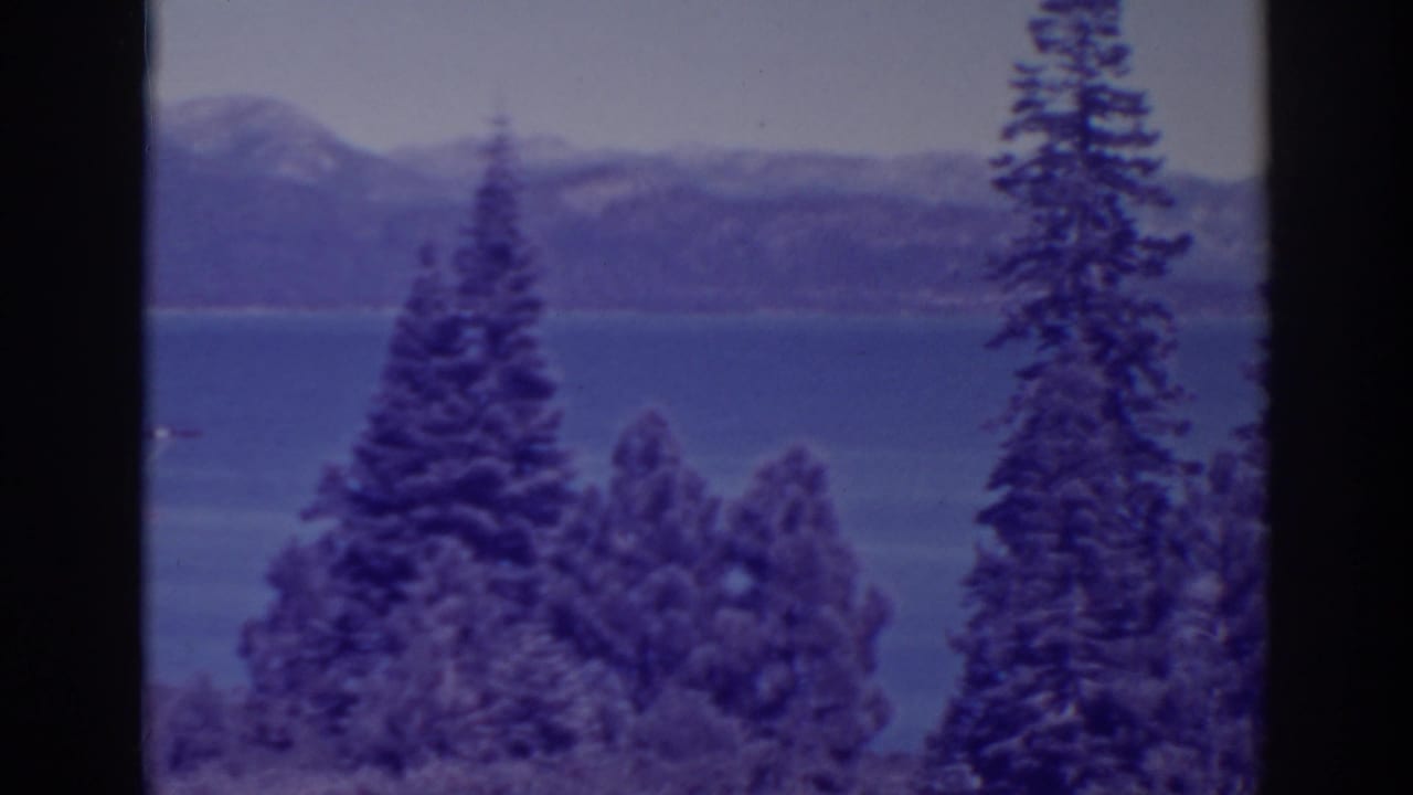 Conifers frame a blue lake and distant mountains in soft-focus 8mm footage.