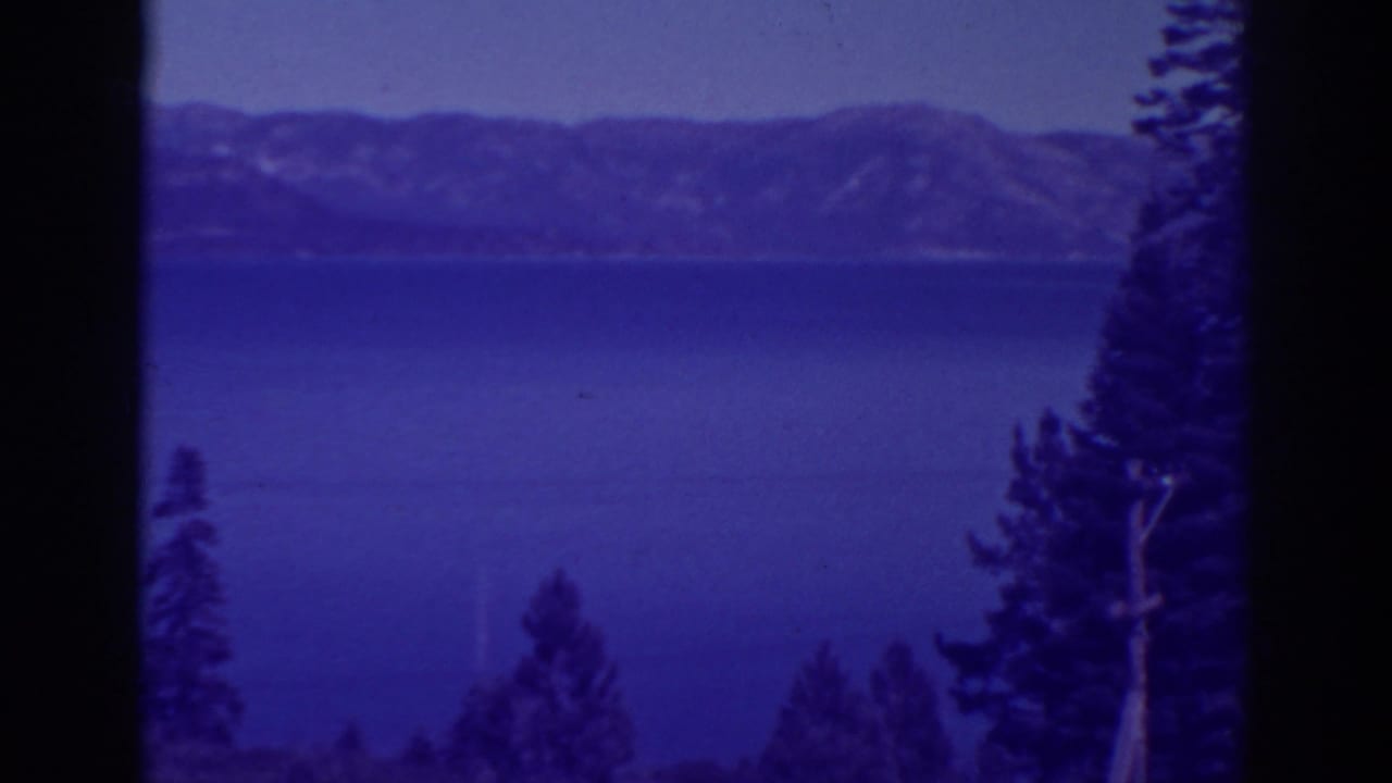 Blue lake and distant mountains seen through dark foreground tree trunks and pines.