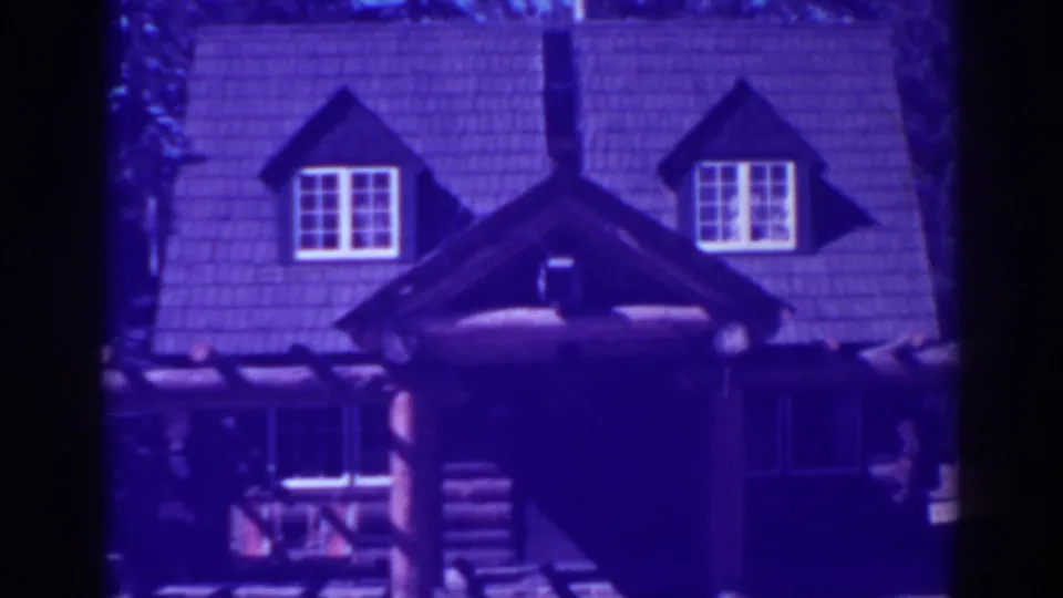 1938 home-movie frame from El Dorado City showing the lodge facade, shingled roof, central chimney, dormer windows, and log porch details.