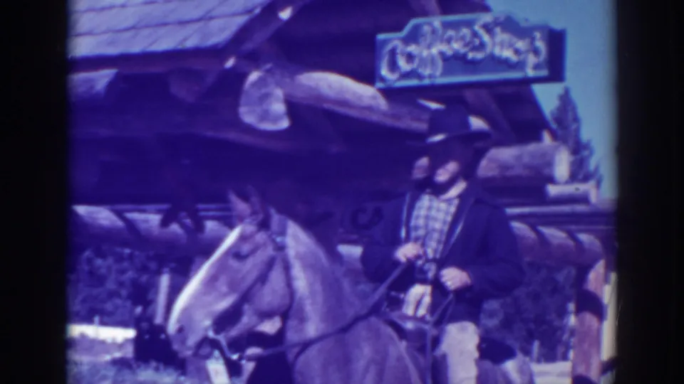 Closer 1938 home-movie frame of a man in a dark hat and jacket seated on a horse near a rustic storefront sign.
