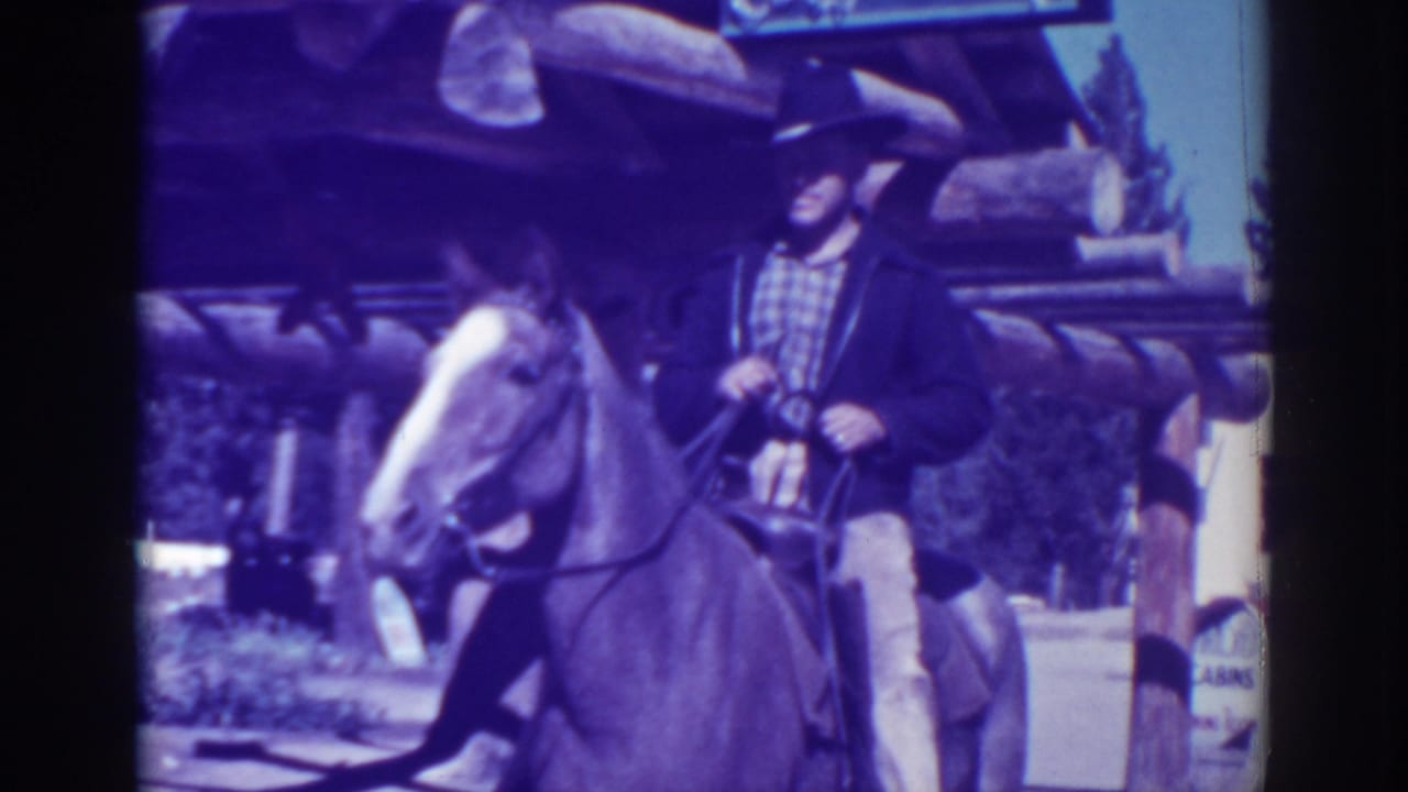 Man in a cowboy hat rides a light-colored horse beside a rustic log cabin structure.