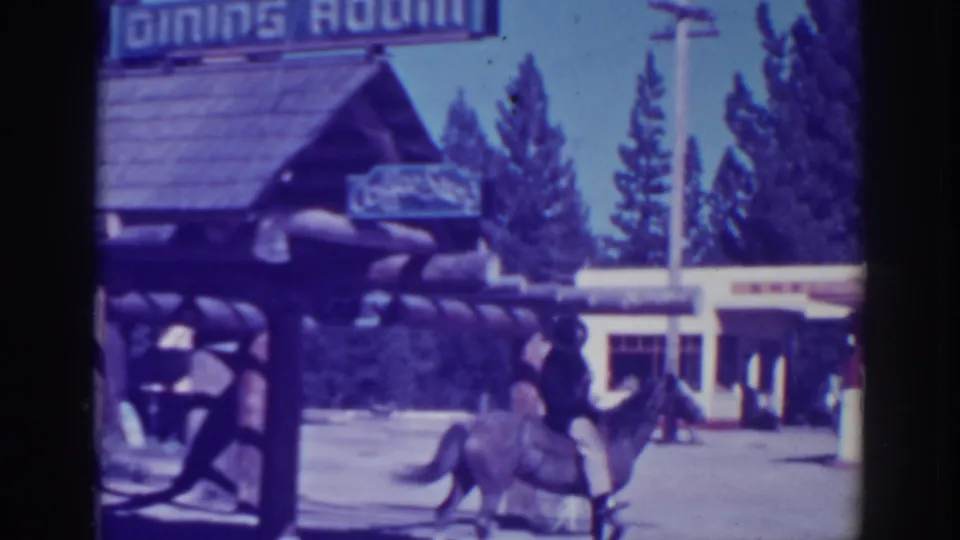 The rider on a light-colored horse passes the rustic dining-room building in El Dorado City, with signage, roadway, and tall trees visible.