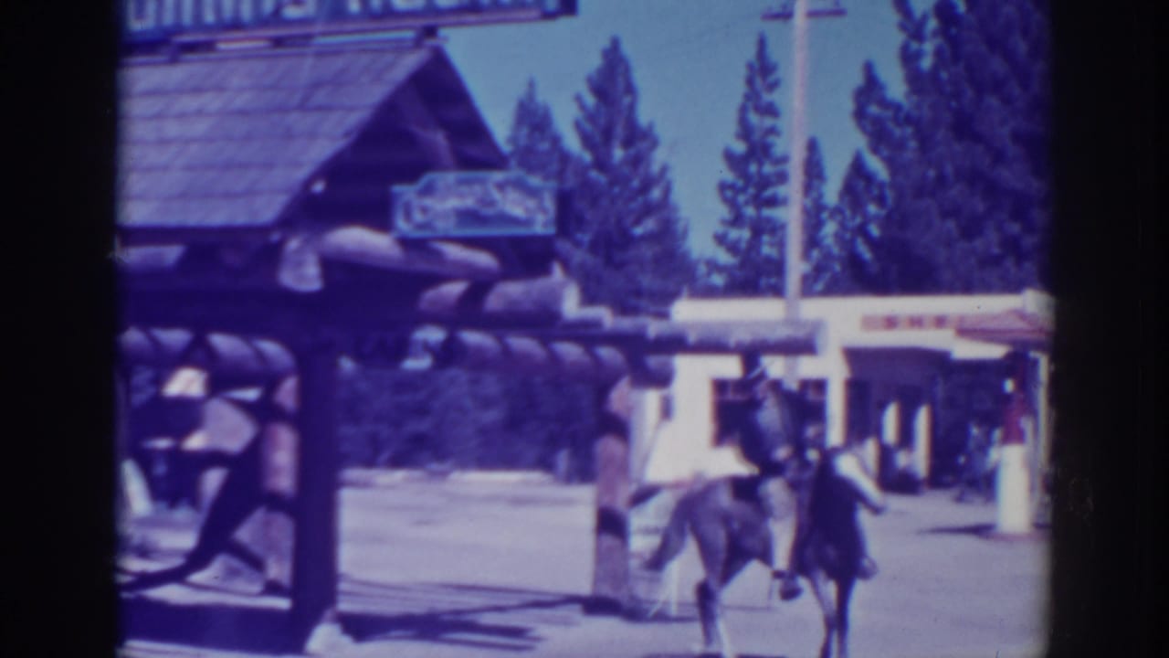 Man on horseback approaches a rustic log gateway with signage and trees behind him.