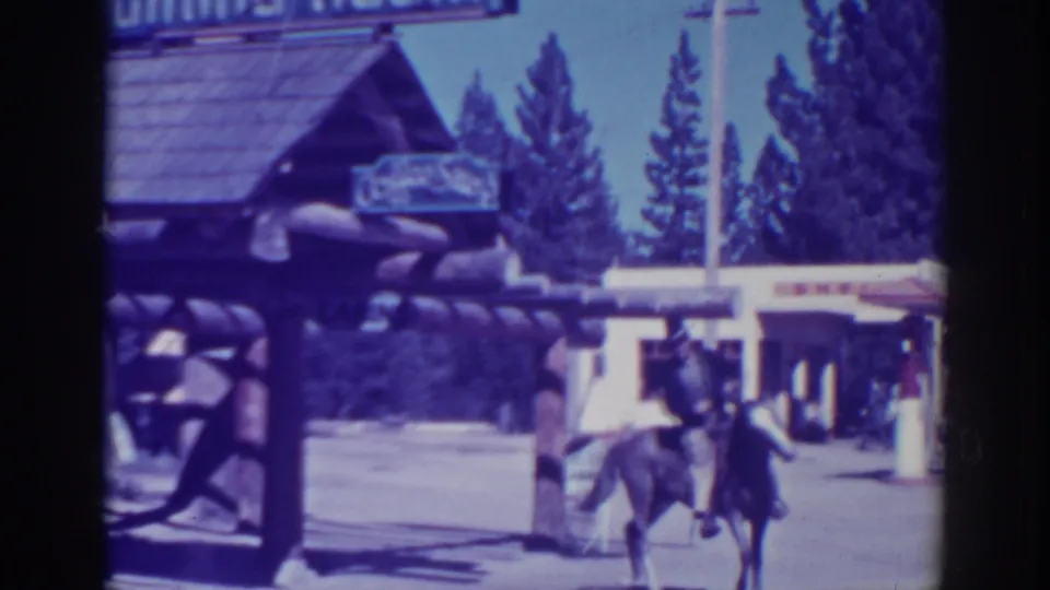 In 1938 El Dorado City, California, a man rides a horse near a rustic log entry structure with signage and tall trees in the background.