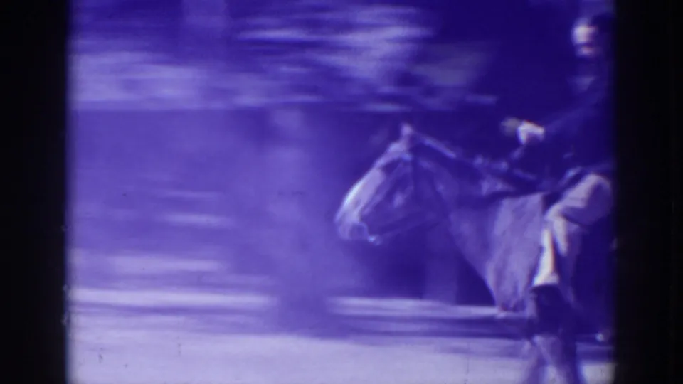 A man in a dark jacket and light pants rides a horse across a dusty path, softened by motion blur and purple 8mm color shift.