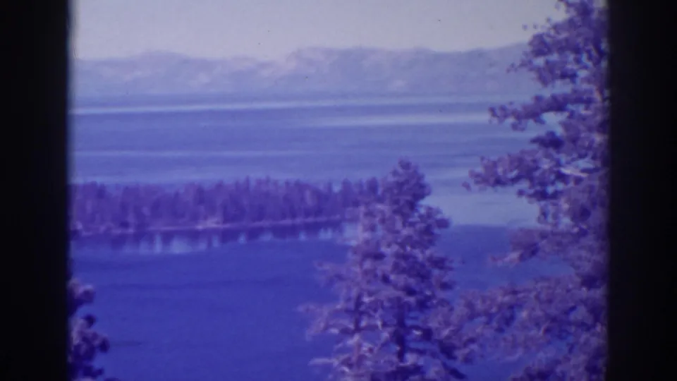 1938 Emerald Bay view opens wider to blue water, a forested shoreline, hazy mountains, and foreground trees.