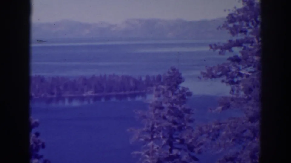 1938 Emerald Bay view with blue lake water, a forested island, distant mountain ridges, and dark evergreen trunks framing the scene.