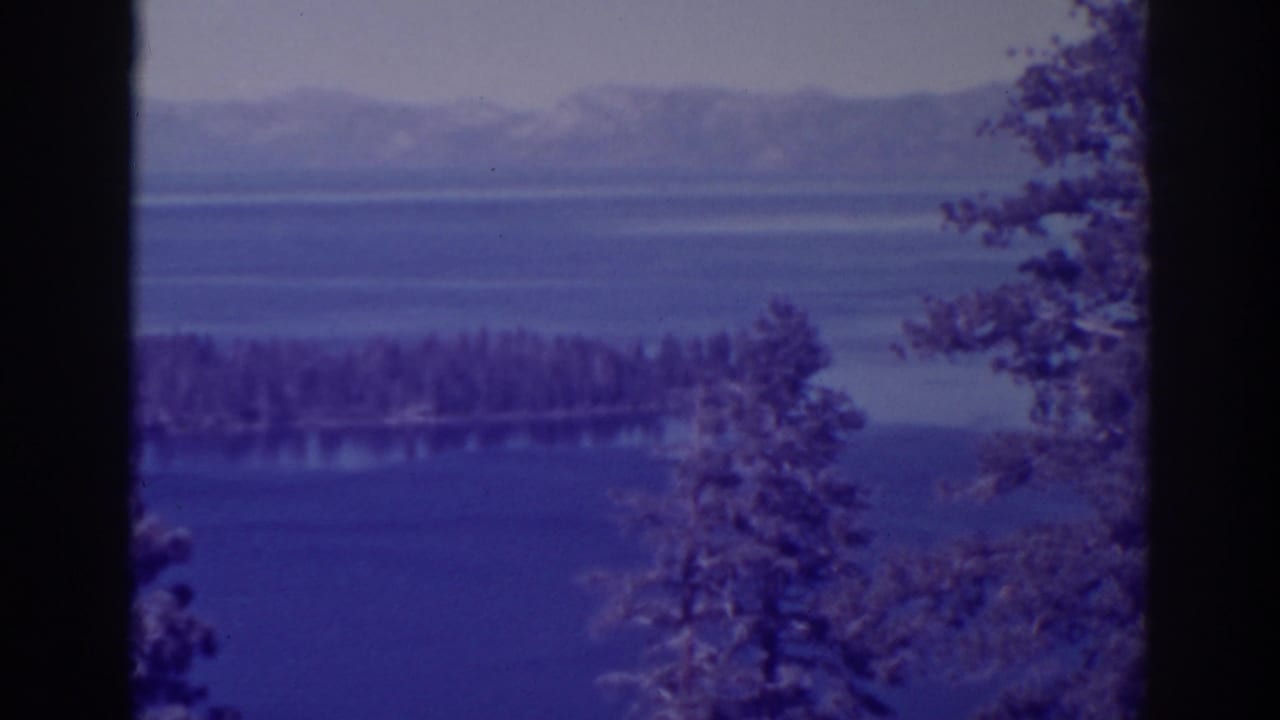 Forested island on a blue lake with distant mountains and evergreen branches at the sides.