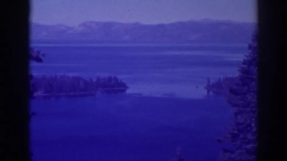 Late frame from the 1938 Emerald Bay clip, with a soft blue lake surface, wooded island, hazy mountain ridge, tree edge, and dark film-frame border.