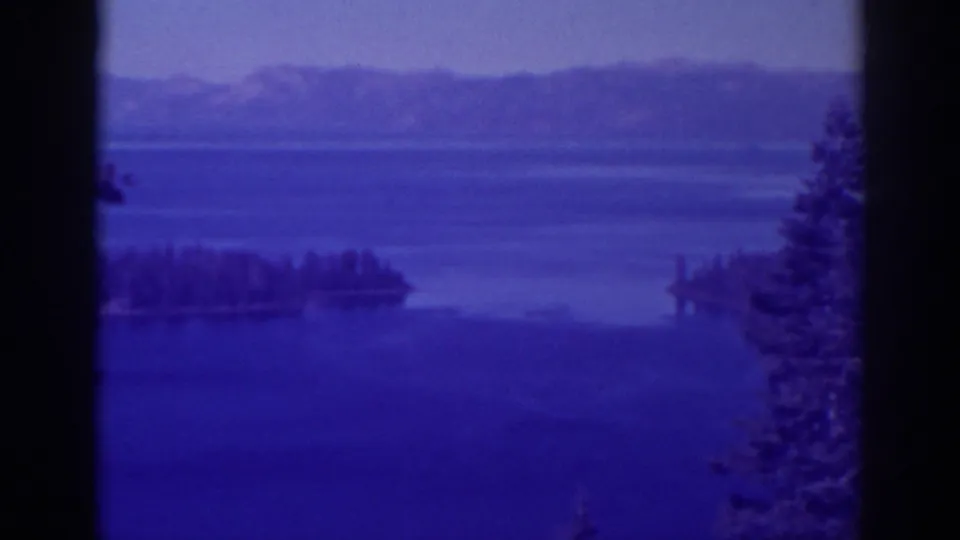 Emerald Bay, California appears in a blue-cast 8mm frame with wooded islands, calm water, and distant mountain ridges.