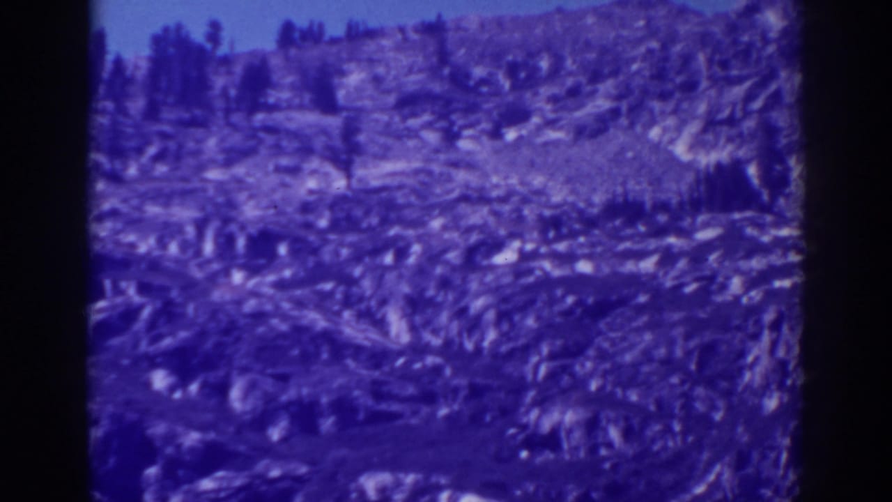 Rocky cliff slope with scattered evergreens and a blue-tinted film border.
