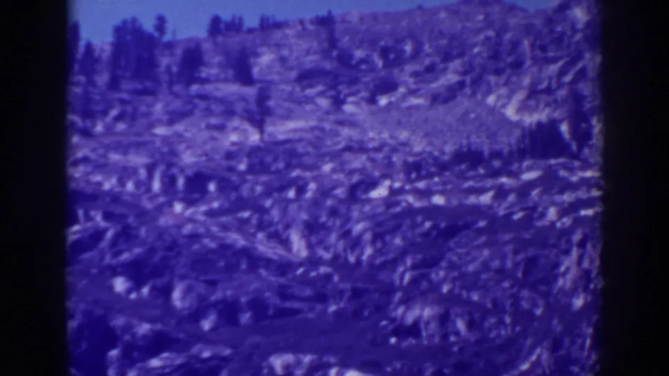 1938 Emerald Bay, California 8mm frame showing a rugged rocky slope, small evergreen trees, and a pale sky with strong blue color shift.