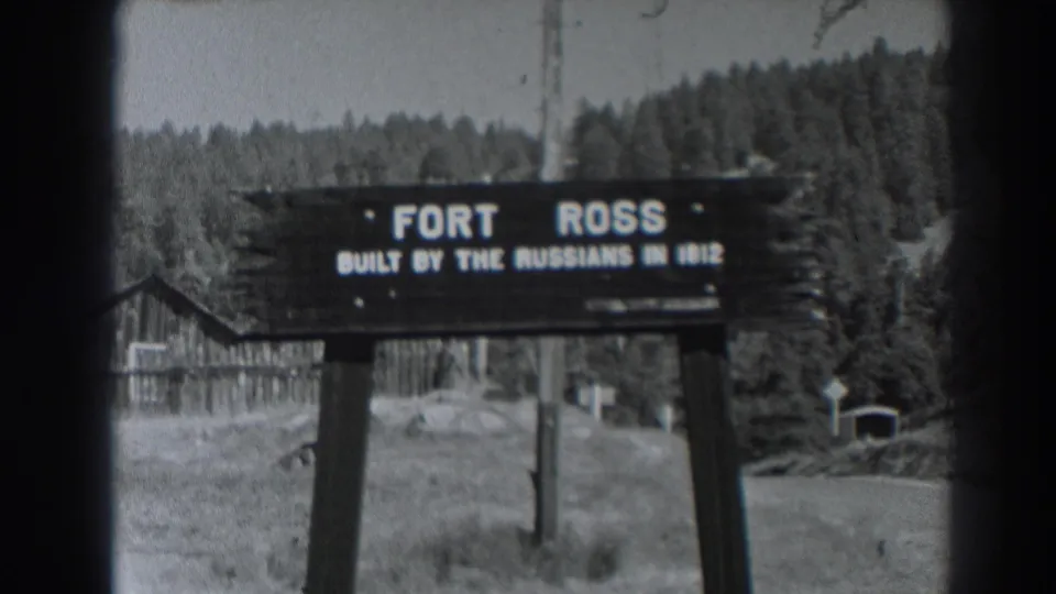1938 Fort Ross, California sign reading built by the Russians in 1812, seen before rustic buildings and a wooded hillside.