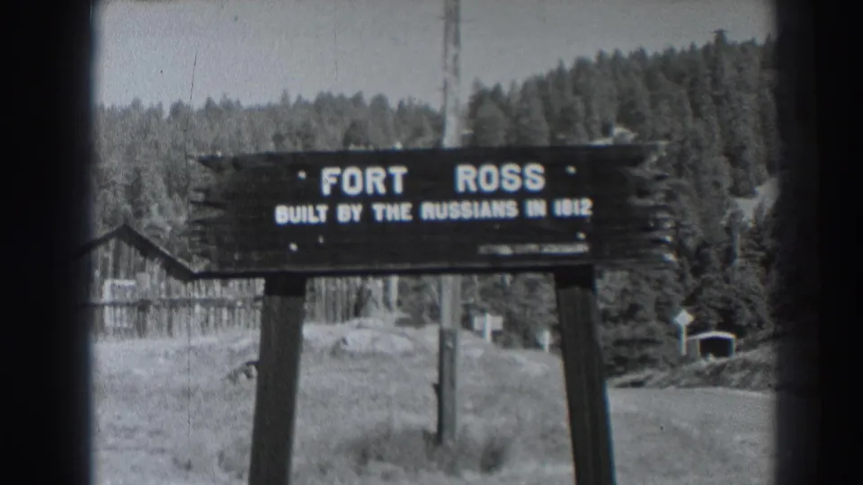 Black-and-white 8mm frame of the Fort Ross, California sign with rustic site buildings and forest visible behind it.