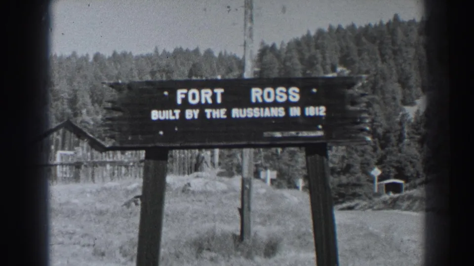 1938 black-and-white 8mm frame showing the Fort Ross sign reading built by the Russians in 1812, with grass, trees and wooden structures behind.