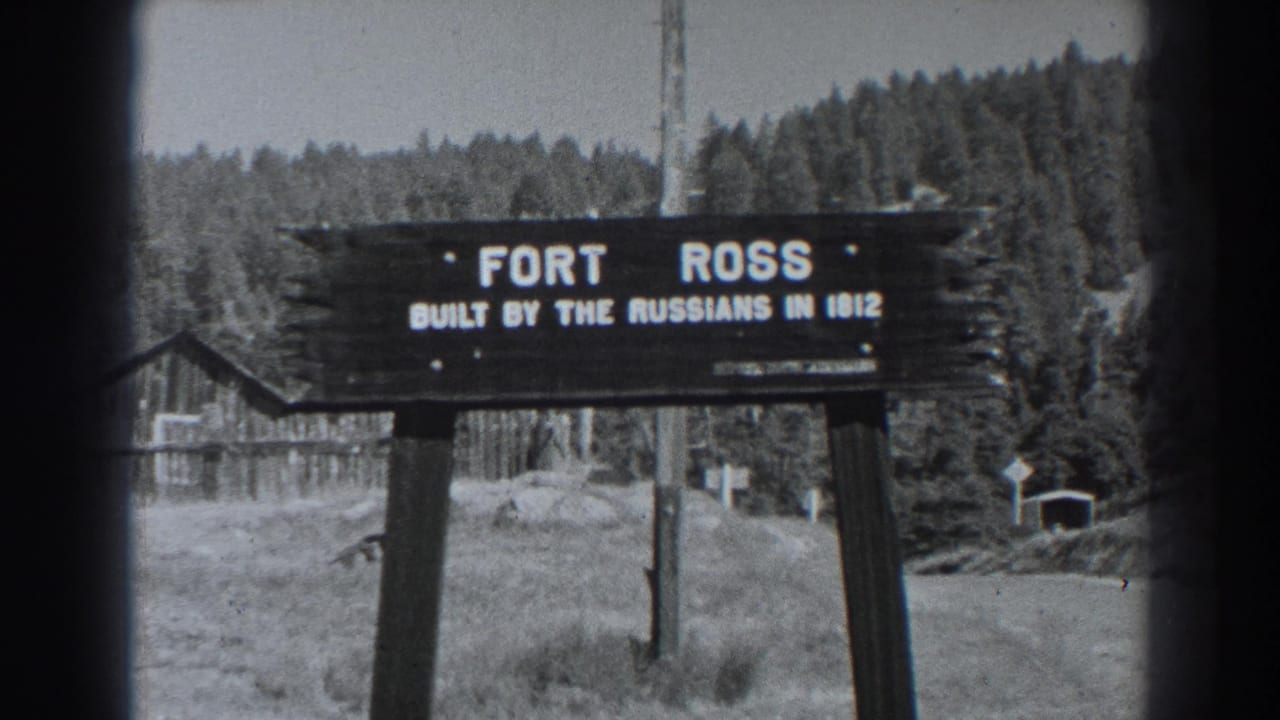 Black-and-white Fort Ross visitor view with the wooden sign centered, dense trees behind it and a small structure near the right edge.
