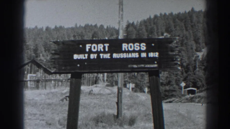 Black-and-white Fort Ross visitor view with the wooden sign centered, dense trees behind it and a small structure near the right edge.