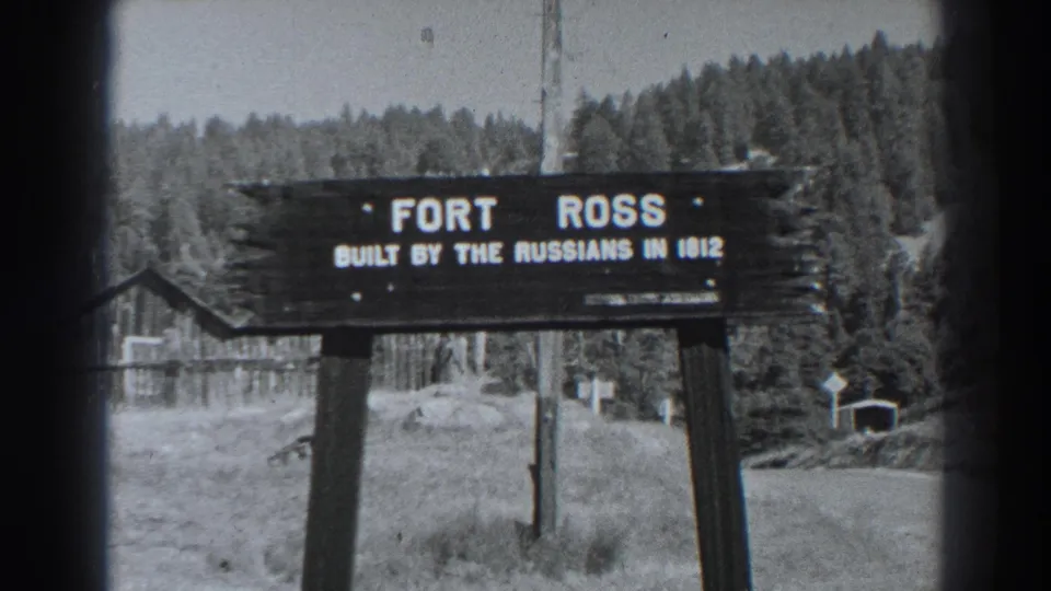 Black-and-white frame of the Fort Ross landmark sign at Fort Ross, California, with wooded hills, grass and simple site structures visible.