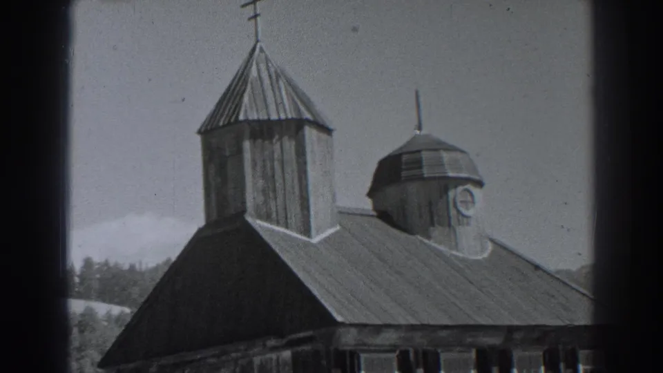 Fort Ross, California frame from 1938 showing a tall wooden tower with cross, domed cupola, sloped roof, treeline, and pale sky.