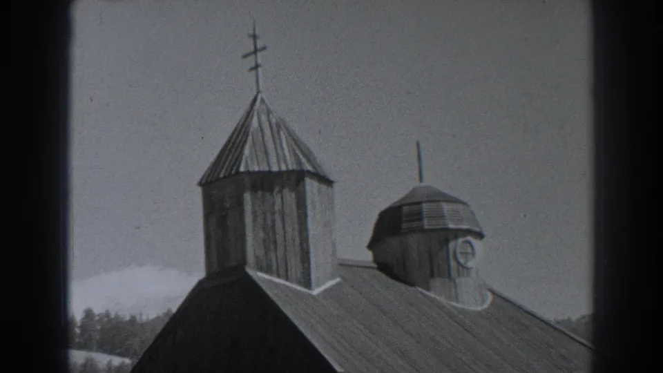 1938 Fort Ross 8mm frame showing the wooden building from a slightly lower angle, with tower, cross, cupola, roof panels, windows, and trees.