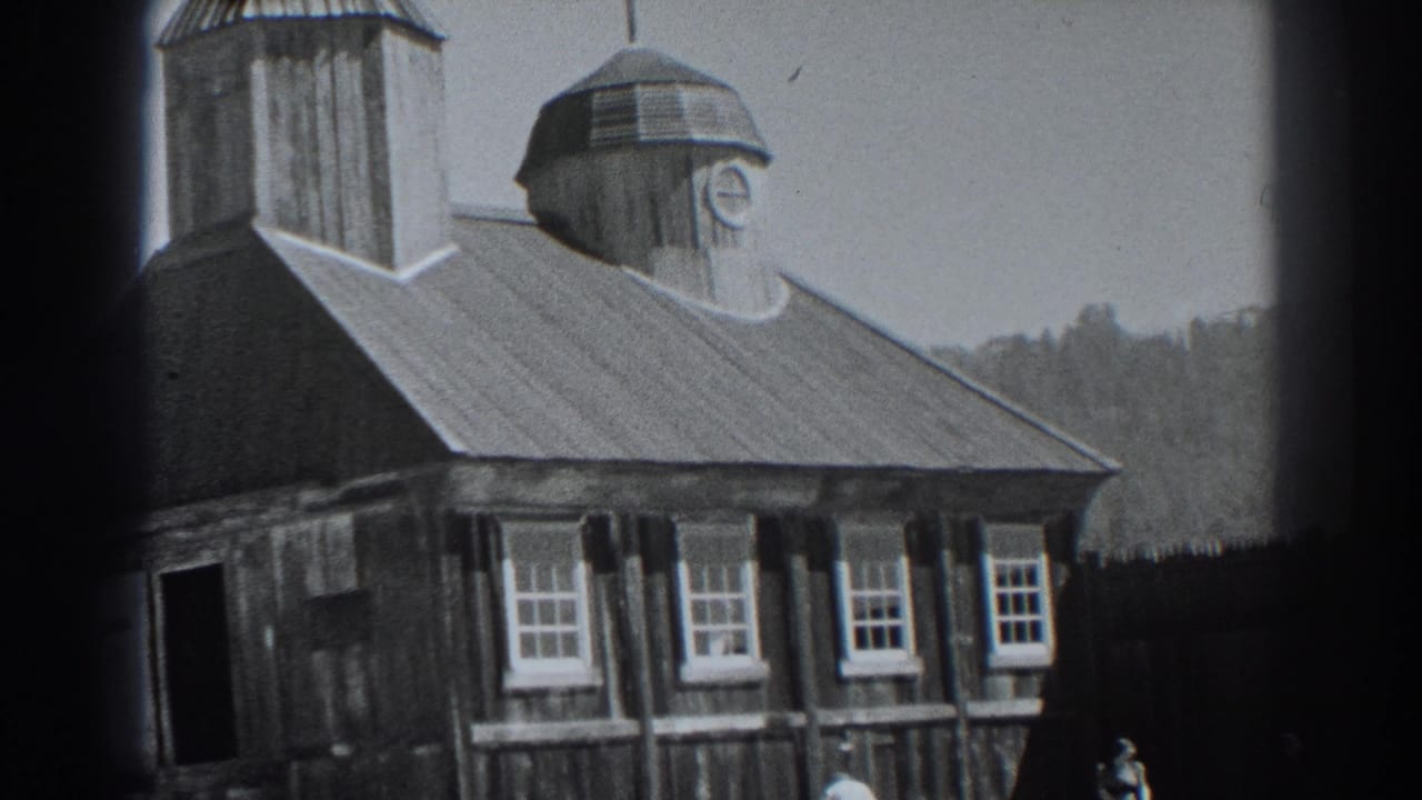 Wooden building with sloped roof, cupola clock, windows, fence, hillside, and small figures.