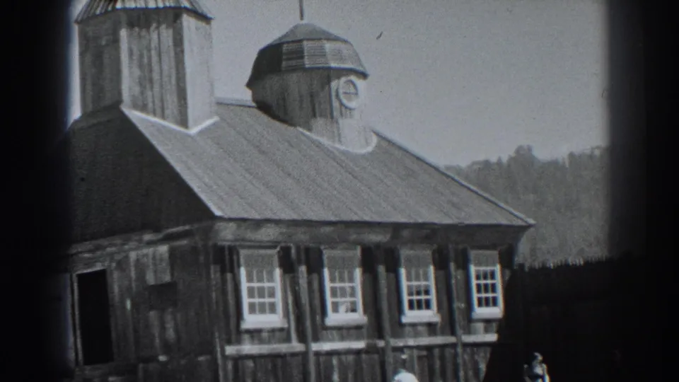 1938 Fort Ross exterior view of a weathered wooden building with a clock-topped cupola, windows, dark fence, hillside, and small people below.
