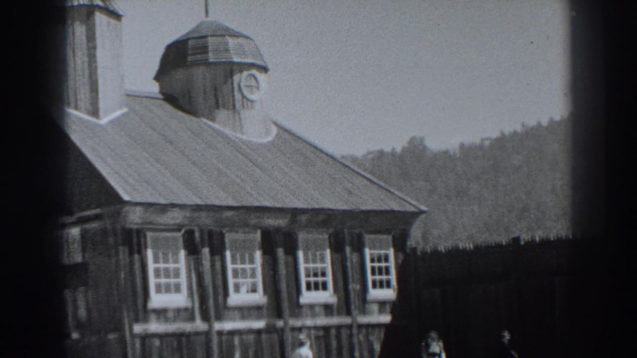 Fort Ross building exterior in 1938, showing the sloped roof, clock cupola, multi-pane windows, dark fence, wooded hill, and indistinct visitors.