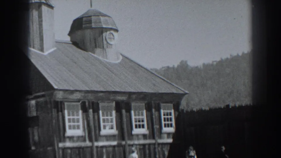 Fort Ross building exterior in 1938, showing the sloped roof, clock cupola, multi-pane windows, dark fence, wooded hill, and indistinct visitors.