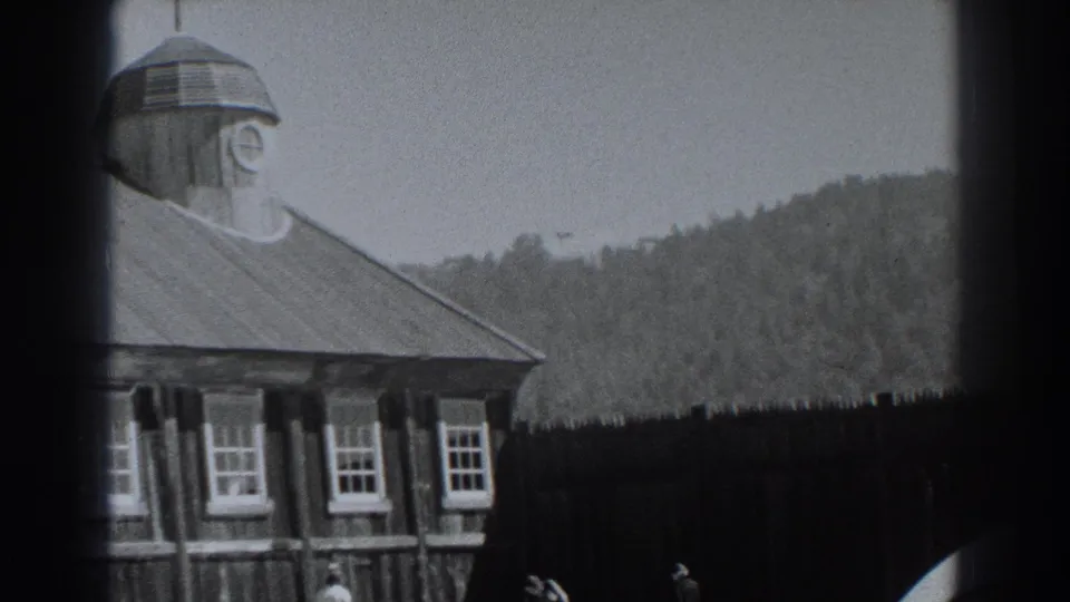 Late shot of the Fort Ross wooden structure, with the clock cupola, sloped roof, window row, dark fence, wooded hillside, and indistinct visitors.
