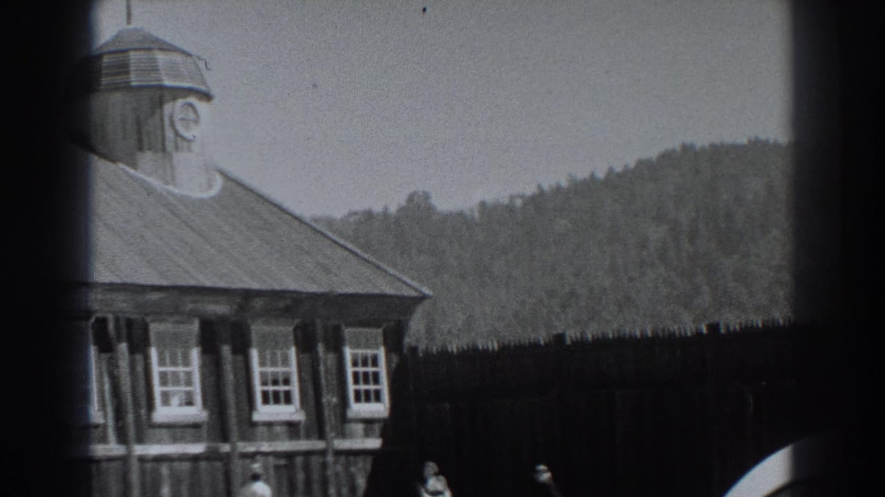 1938 Fort Ross home-movie frame showing a wooden historic building with cupola, white-trimmed windows, wooded hill, and small distant visitors.