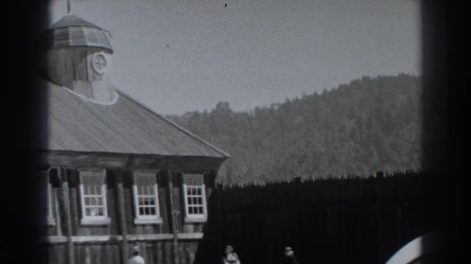 1938 Fort Ross home-movie frame showing a wooden historic building with cupola, white-trimmed windows, wooded hill, and small distant visitors.