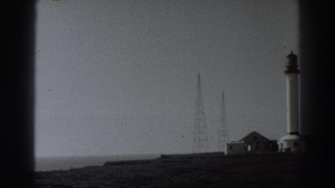 Fort Ross area view with a tall coastal tower, radio masts, low buildings, open ground, and the soft contrast of 1938 black-and-white 8mm film.